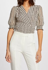 Blusa beige transparente con estampado geométrico negro, escote en V, mangas abullonadas gatheradas y puños elásticos, combinada con pantalones blancos de corte ajustado.