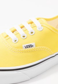 Žluté plátěné tenisky s bílými tkaničkami, černobílou pruhovanou podrážkou a malou štítkovou značkou Vans na straně blízko tkaniček.