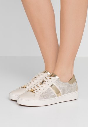 Voeten met lage sneakers in crème met gouden metallic accenten en fonkelende zijpanelen, staand op een lichtgrijze ondergrond.
