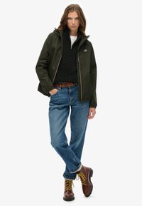 Wodoodporna kurtka w kolorze oliwkowej zieleni z zamkiem z przodu, zestawiona z czarnym swetrem w sploty i luźnymi niebieskimi dżinsami. Brązowe sznurowane buty.