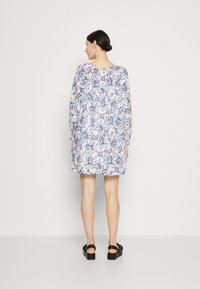Blomsterkjole med lange ermer, hvit bunn, blå og lilla bladmønster, avslappet passform, over kneet lengde, kombinert med svarte platåsandaler.