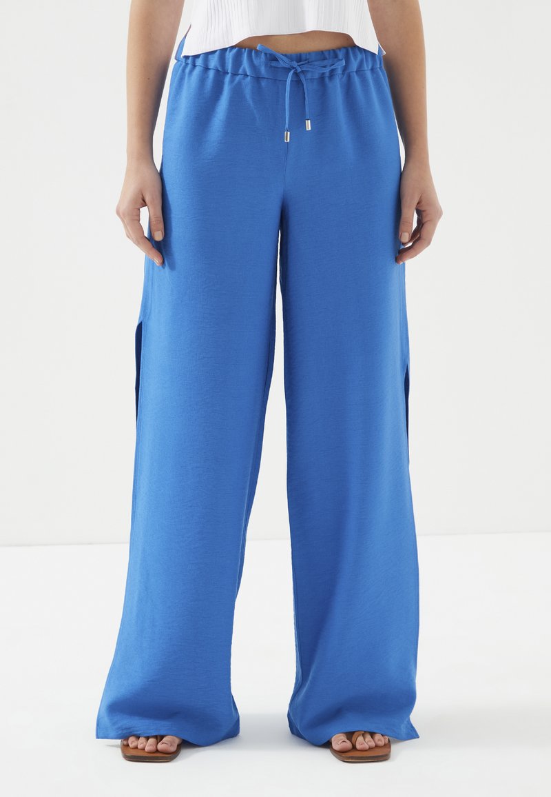 Blauwe, brede broek van zachte stof met een elastische tailleband en trekkoord. Bevat zijslitsen en een soepele textuur.