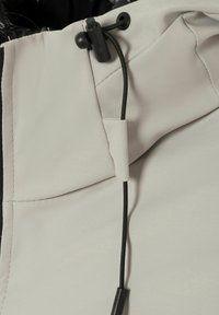 Beige kledingstuk met een zwarte verstelbare trekkoord met een zwarte plastic toggle om de pasvorm aan te spannen of losser te maken.