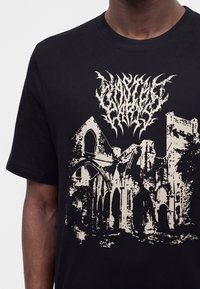 Μαύρο T-shirt με μπεζ γραφικό παλαιών πέτρινων αψίδων και πύργων, που κορυφώνονται με ένα περίπλοκο, δυσανάγνωστο στυλιζαρισμένο κείμενο.