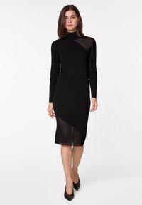 Robe en tricot noire à manches longues et col haut, présentant un design asymétrique avec une section inférieure transparente et des détails côtelés.