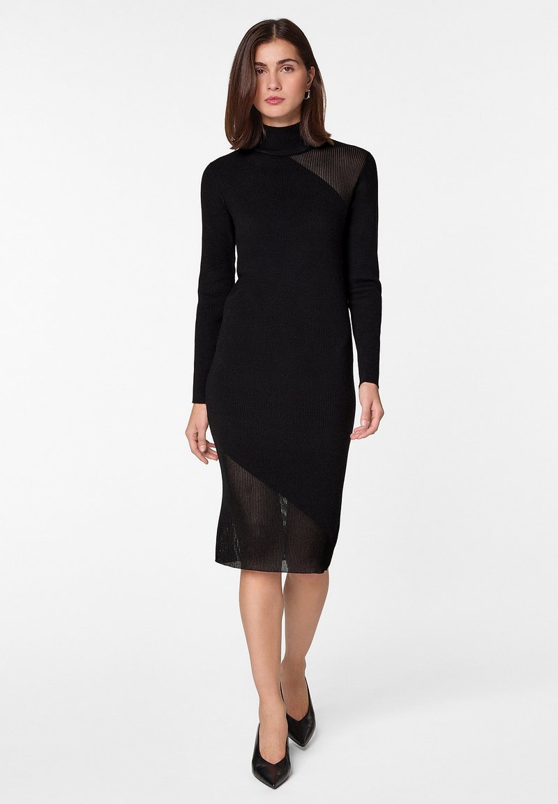 Robe en tricot noire à manches longues et col haut, présentant un design asymétrique avec une section inférieure transparente et des détails côtelés.
