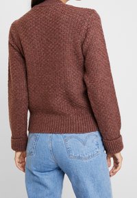 Femme portant un pull en maille texturée couleur rouille et un jean taille haute bleu clair, vue de dos sur un fond uni.