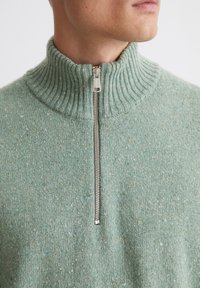 Hellgrüner Strickpullover mit hohem, geripptem Kragen und einem silbernen Reißverschluss. Die Textur weist Sprenkel aus dunkleren Garnen auf.