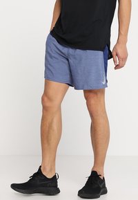 Blå atletiska shorts för män i lättviktsmaterial, med en texturerad yta och en vit logotyp på sidan. I kombination med svarta sneakers.