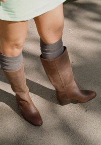 Botas marrones de cuero hasta la rodilla con punta afilada y un leve tacón cuadrado, combinadas con calcetines de punto grises por encima de la pantorrilla, sobre una superficie de concreto.