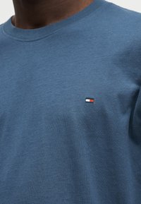 Άνδρας που φοράει ένα απλό μπλε μπλουζάκι με στρογγυλή λαιμόκοψη και ένα μικρό ορθογώνιο λογότυπο σε κόκκινο, λευκό και ναυτικό μπλε στο αριστερό στήθος.