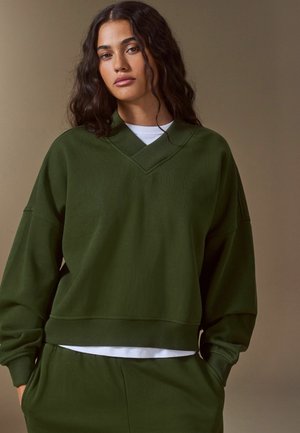 Kvinde med langt mørkt hår iført en grøn oversized sweatshirt og matchende bukser, stående med hænderne i lommerne mod en ensfarvet baggrund.