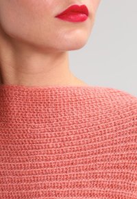 Pull tricoté couleur pêche avec un motif texturé et ample. Encolure légèrement décontractée et finition douce et duveteuse, rehaussée par des lèvres rouge vif.