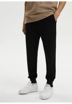 Mężczyzna w czarnych spodniach dresowych i białych trampkach, stojący z jedną ręką w kieszeni na jednolitym białym tle.