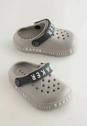 Zoccoli di colore beige chiaro realizzati in materiale sintetico con numerosi fori di ventilazione. Presentano una fascia nera con il marchio "BAKER" e un dettaglio del logo.