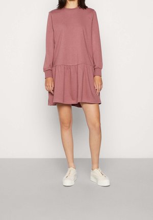 Une femme se tient debout portant une robe rose poussiéreuse à manches longues, coupe ample avec une taille froncée, et des baskets blanches à plateforme sur un fond uni.