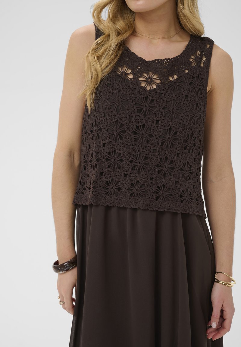 Femme portant un haut sans manches en dentelle fleurie en crochet noir sur une robe noire assortie, avec des bracelets aux deux poignets.