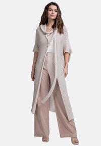 Beżowy sweter z kapturem, krótki rękaw, otwarty przód i związywane zapięcie. Połączony z jasnymi spodniami o szerokich nogawkach i przezroczystymi sandałami.