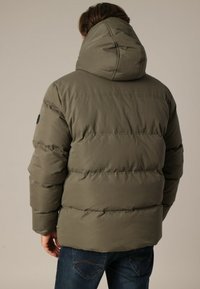 Doudoune verte olive avec capuche, présentant un design matelassé, des poignets de manche élastiques et un patch circulaire noir sur le bras gauche.
