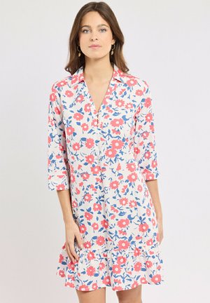 Femme portant une robe blanche à imprimé floral rouge et bleu, manches trois-quarts, et ourlet à volants, se tenant devant un fond clair uni.