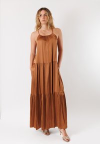 Abito maxi marrone realizzato in materiale lucido con spalline sottili, corpetto arricciato e gonna a strati. Dotato di tasche laterali e un design fluido.