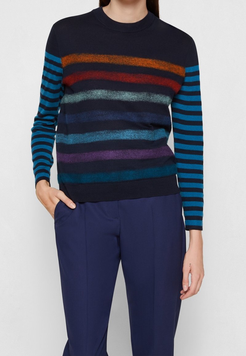 Femme portant un pull bleu marine à rayures horizontales multicolores et manches rayées bleues, associé à un pantalon bleu marine, main dans la poche.