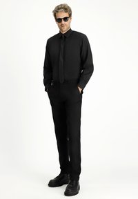 Traje formal negro que incluye una camisa de manga larga y corbata, con pantalones ajustados. El modelo lleva gafas de sol negras y zapatos negros robustos.