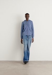 Haut à capuche zippé bleu clair, associé à un jean bleu délavé. Le modèle se tient contre un fond blanc uni, mettant en avant une tenue décontractée.