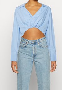 Jasnoniebieska krótka bluzka o fasonie kopertowym, z długimi rękawami i dekoltem w serek; zestawiona z wysokim stanem jasnoniebieskimi dżinsami.