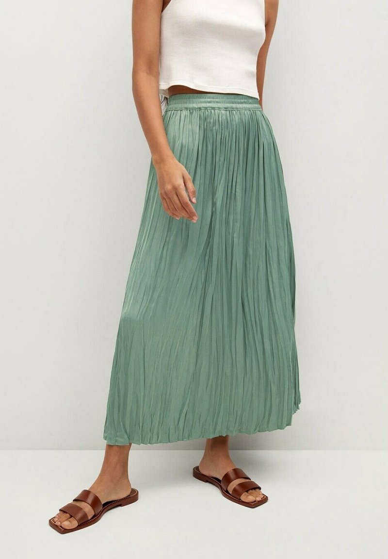 Femme portant un haut blanc court sans manches, une jupe maxi plissée verte taille haute, et des sandales marron à enfiler, debout sur un sol blanc.