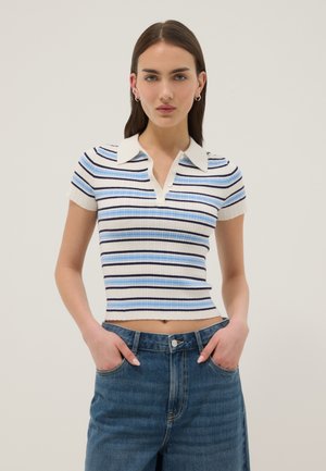 Young woman wearing a short-sleeve, ribbed, striped polo shirt in blue, white, and black paired with high-waisted blue jeans, hands in pockets.