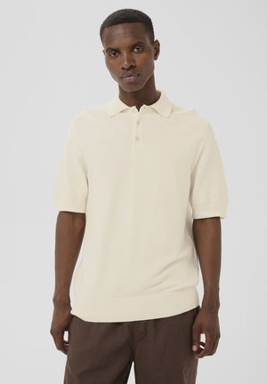Man wearing a cream short-sleeve knit polo shirt and brown pants, standing against a plain light gray background.