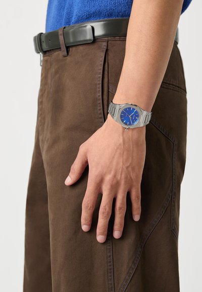 Silver wristwatch with a blue dial and stainless steel band, worn on a hand alongside brown pants with detailed stitching and a black belt.