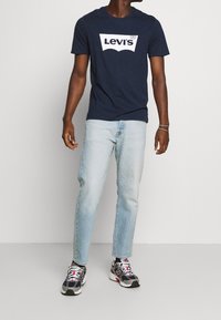 T-shirt blu navy con logo bianco di Levi's, abbinato a jeans azzurri e scarpe da ginnastica grigie con accenti rossi e neri.