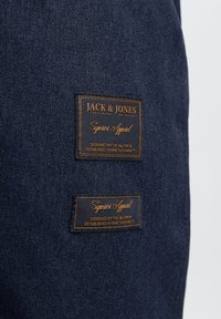 Marineblauer Denim-Stoff mit zwei rechteckigen Etiketten mit goldener Schrift: "JACK & JONES" und "Superior Apparel" auf einem strukturierten Hintergrund.