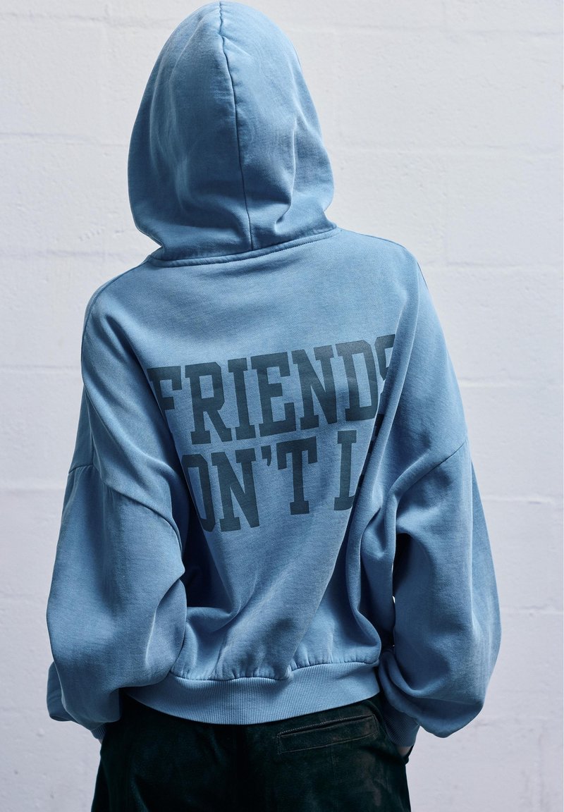 Kék kapucnis pulóver, oversized ujjal. A hátsó részen fekete szöveg olvasható: "A BARÁTOK NEM HAZUDNAK." Puha, laza anyagú textúra.