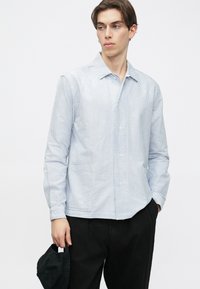 Gestreept overhemd met knopen in lichtblauw en wit, voorzien van een kraag, lange mouwen en twee voorzakken. Draagt bij donkere broek.