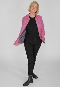 Roze neplederen jas met een kraag, open voorkant en grijze voering, gecombineerd met een zwarte blouse en broek, afgewerkt met zwarte enkellaarsjes.