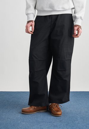 Μαύρο παντελόνι με φαρδύ πόδι και πολλαπλές τσέπες, κατασκευασμένο από ανθεκτικό ύφασμα. Συνδυασμένο με καφέ παπούτσια τύπου boat. Ορατή υφή στην επιφάνεια.