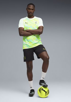 Atleta masculino de pie con los brazos cruzados, un pie sobre un balón de fútbol Puma de color amarillo neón, vistiendo ropa deportiva y tacos Puma, sobre un fondo gris liso.