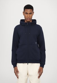 Navy spalvos zip-up džemperis, pagamintas iš minkštos medžiagos, su reguliuojamu gobtuvu, su priekinėmis kišenėmis ir subtiliu logotipo detaliu, derinamas su šviesiai kreminėmis kelnėmis.