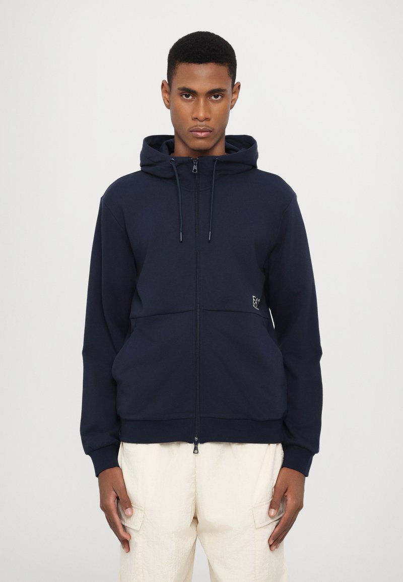 Navy spalvos zip-up džemperis, pagamintas iš minkštos medžiagos, su reguliuojamu gobtuvu, su priekinėmis kišenėmis ir subtiliu logotipo detaliu, derinamas su šviesiai kreminėmis kelnėmis.