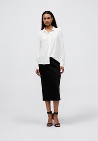 Chemise blanche à manches longues, associée à une jupe midi noire. Le mannequin porte des sandales à talons noires. Tenue simple et professionnelle.