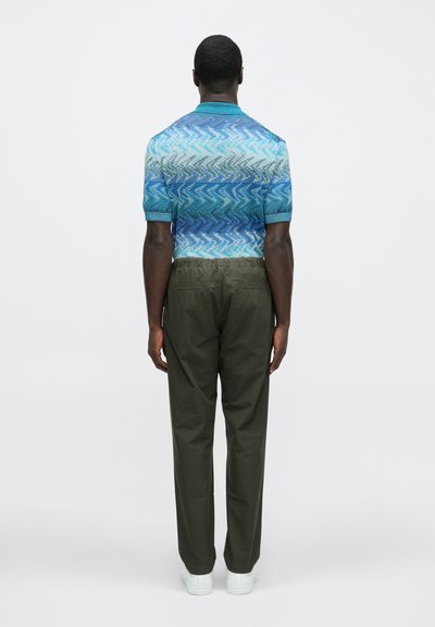 Πλεκτό πουκάμισο με κοντά μανίκια και διαβάθμιση μπλε μοτίβου· συνδυασμένο με παντελόνι ελιάς και λευκά αθλητικά παπούτσια, εμφανιζόμενο από πίσω.