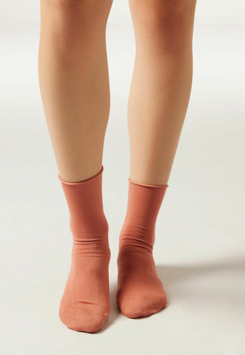 Calcetines de tobillo acanalados de color rosa claro, diseño ajustado, textura suave, con punta redondeada y parte superior elástica, visibles sobre las piernas contra un fondo neutro.