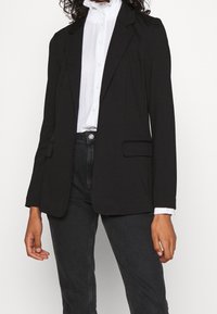 Musta bleiseri, jossa on lovelliset kaulukset, yksittäinen napitus ja kaksi etutaskua. Päällä valkoinen korkeakauluksinen paita ja yhdistetty tummiin farkkuihin.