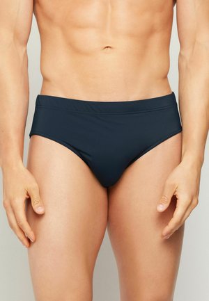 Männlicher Oberkörper trägt dunkelblaue Badehose, steht mit entspannten Armen seitlich vor neutralem Hintergrund.