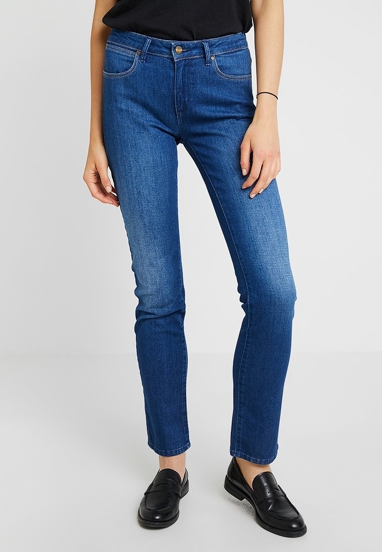 Vaqueros skinny de color azul oscuro con una cintura de media altura, ligeras decoloraciones, bolsillos frontales y detalles de costura visibles, combinados con mocasines negros.