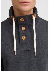 Bluză gri cu guler înalt, având patru butoni maro și șireturi albe contrastante. Material din bumbac texturat.