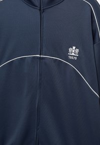 Námořnicky modrá bunda na zip s bílým lemováním přes hrudník a ramena, s malým vyšívaným logem věže s květinami a číslem 15573.